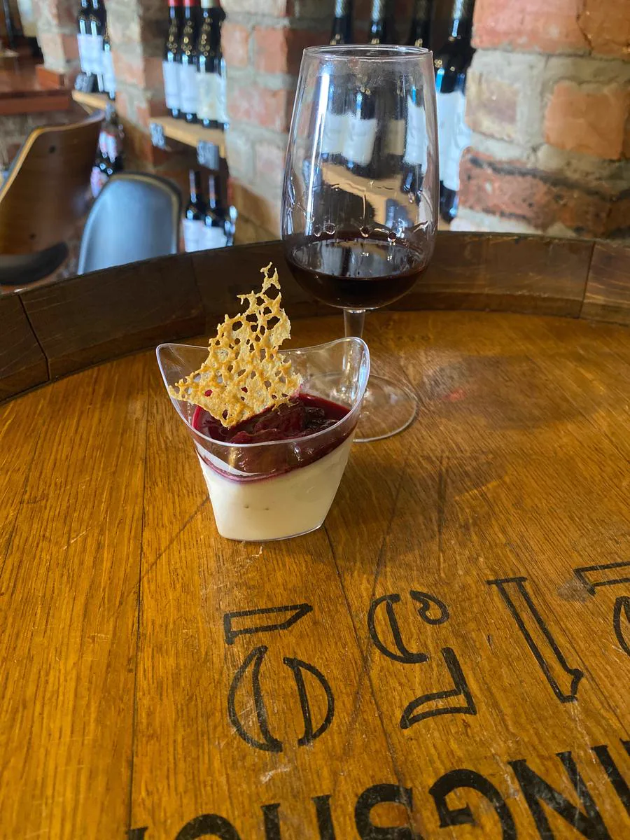 A glass of red wine and a plated dessert on an oak barrel head, bottle racks and a brick wall behind